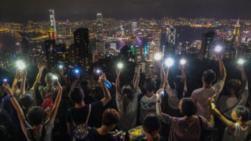 A view from behind of a group of people standing and overlooking the Hong Kong skyline at night. The people hold with their phones high with the flashlight turned on.